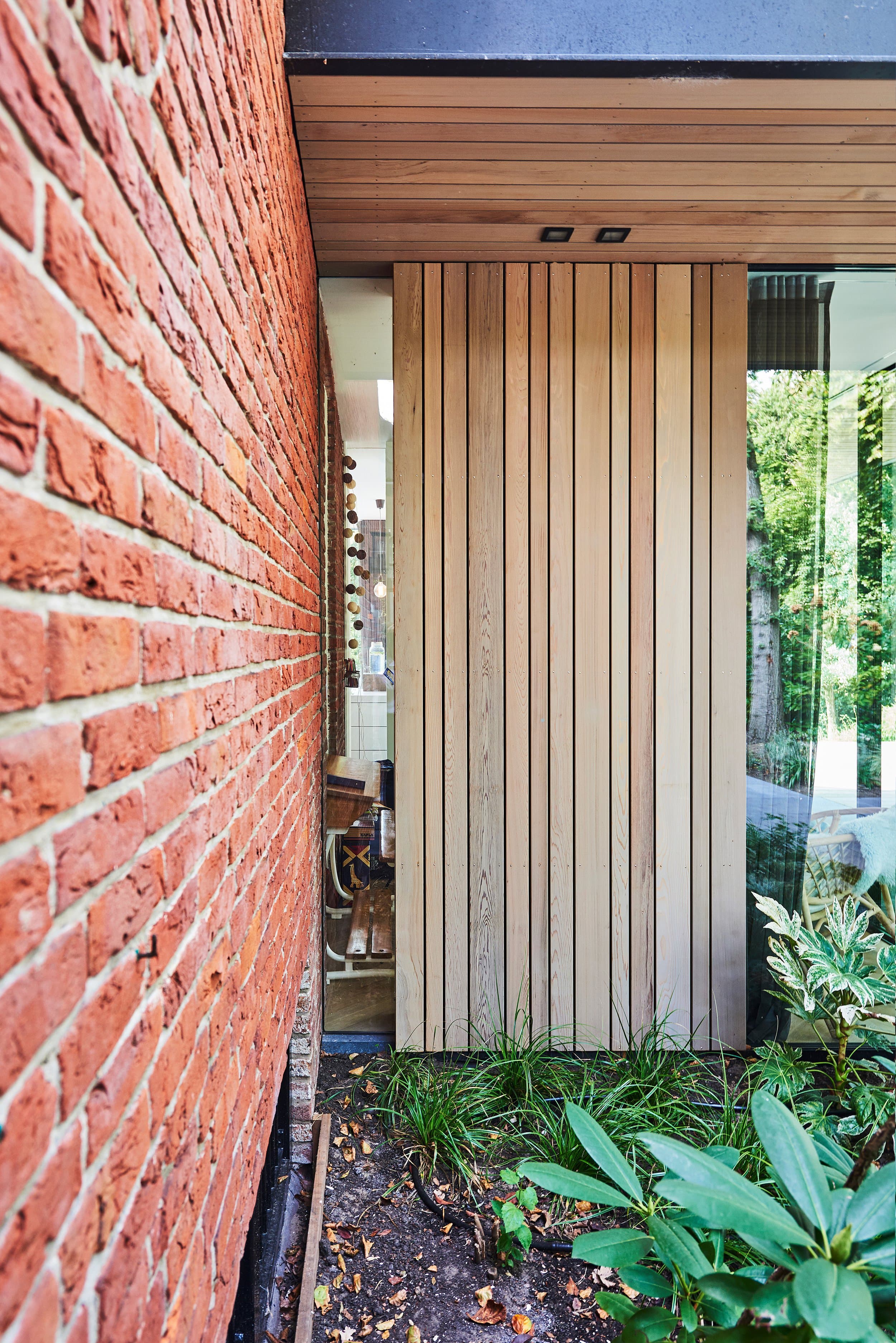 Glazen deur met verticale houten lamellen, bakstenen muur links zichtbaar