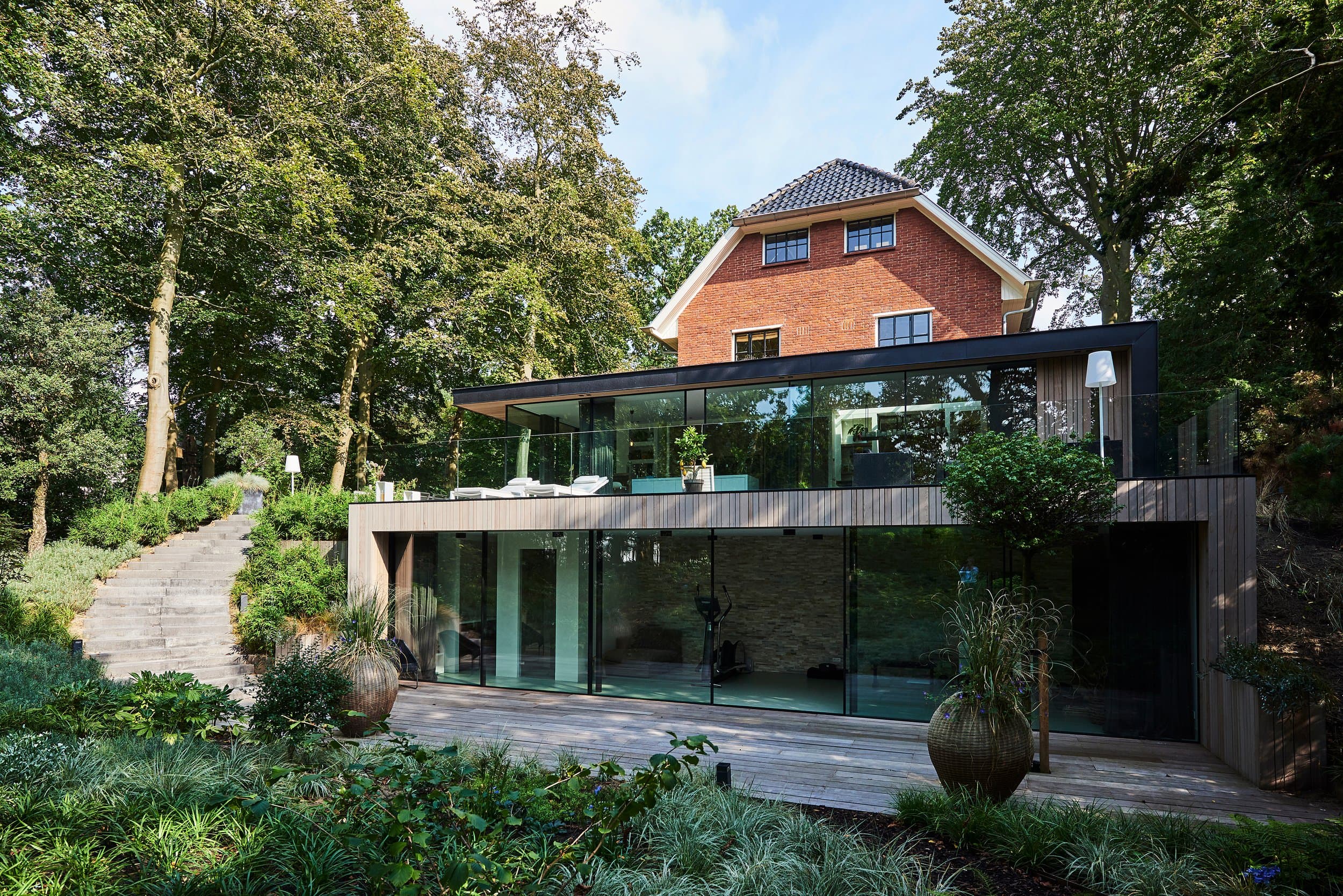 Moderne uitbouw met glas, zwart stalen frame en houten terras aan bakstenen villa