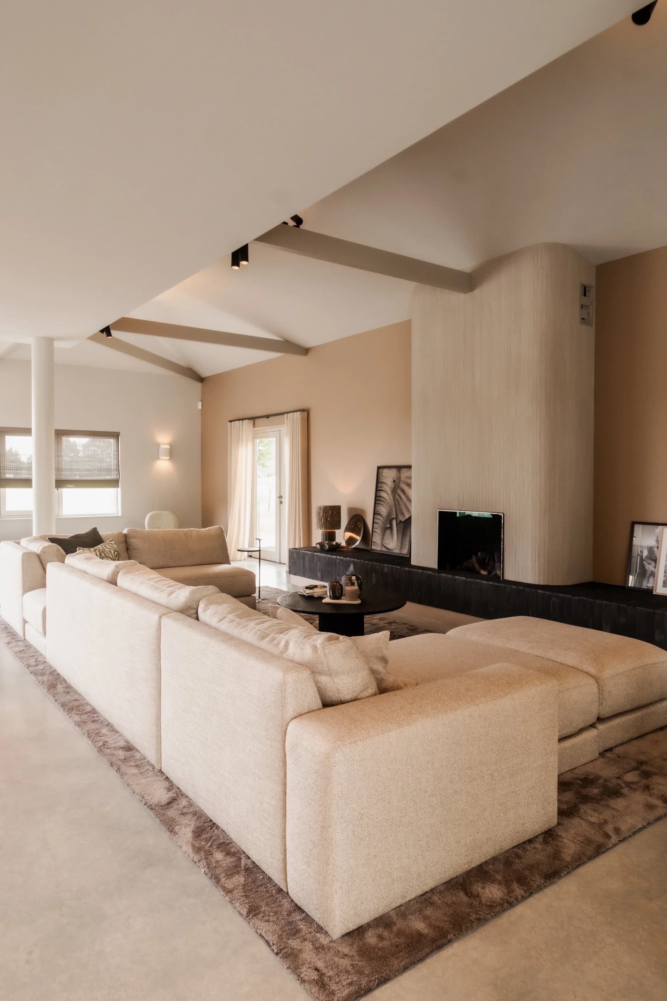 Woonkamer met beige hoekbank en zwarte haard, houten balken