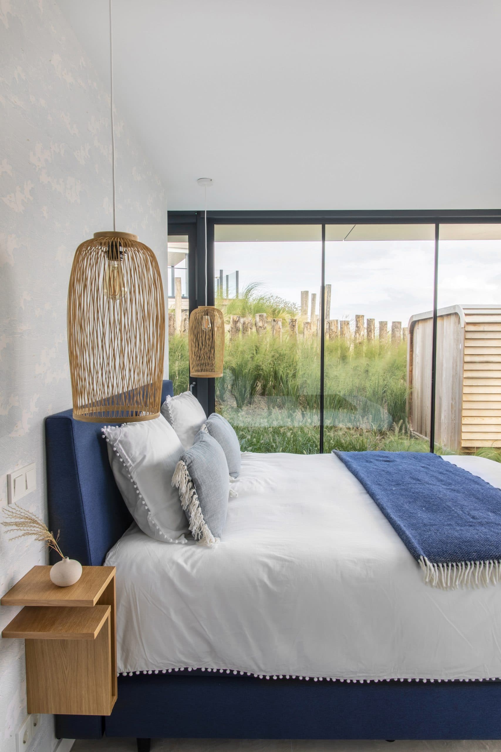 Slaapkamer met donkerblauwe boxspringbed, rieten hangende lamp en watertuin uitzicht