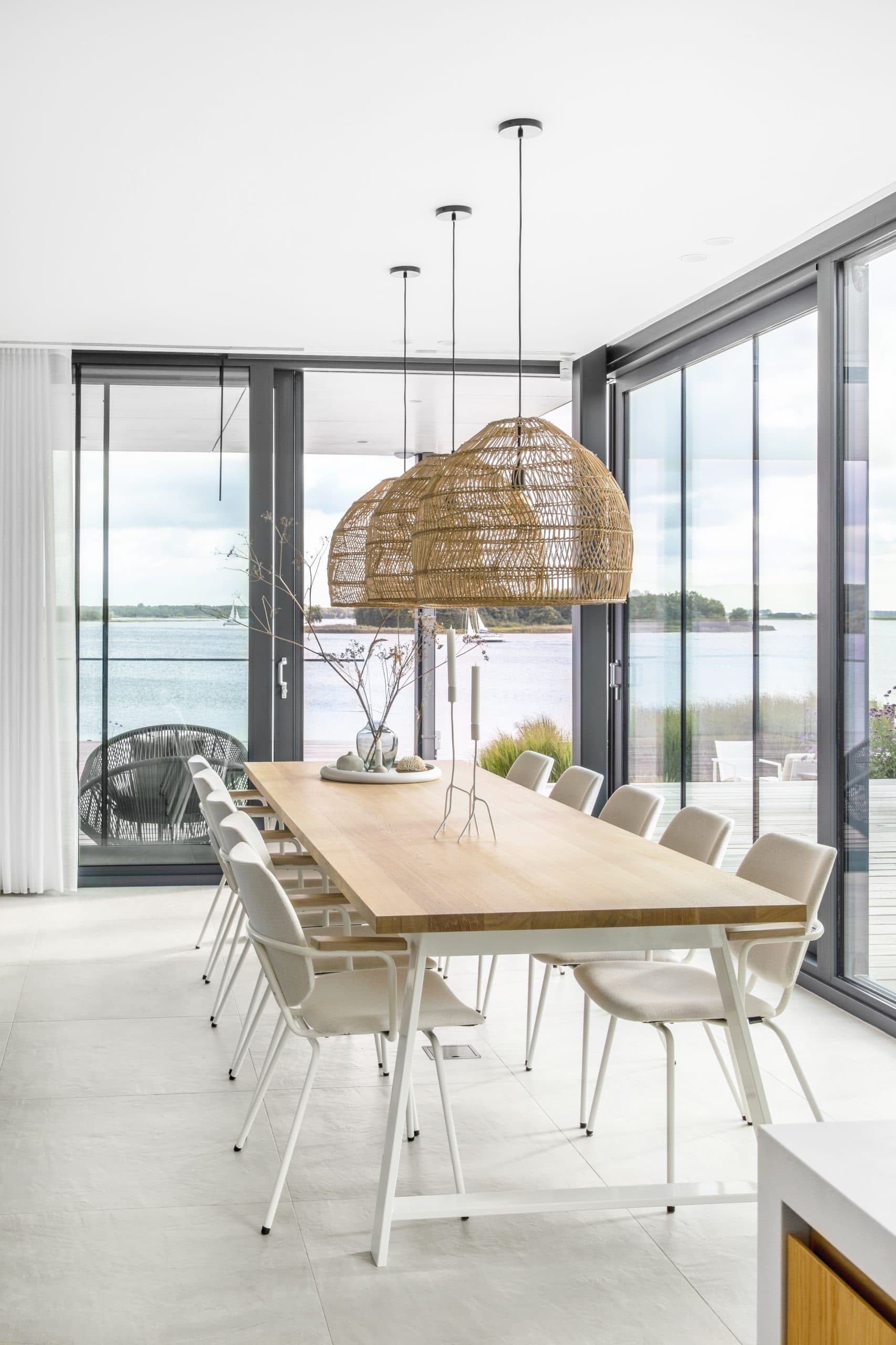 Diningzone met houten tafel, rieten hanglamp en wateruitzicht door glazenwand