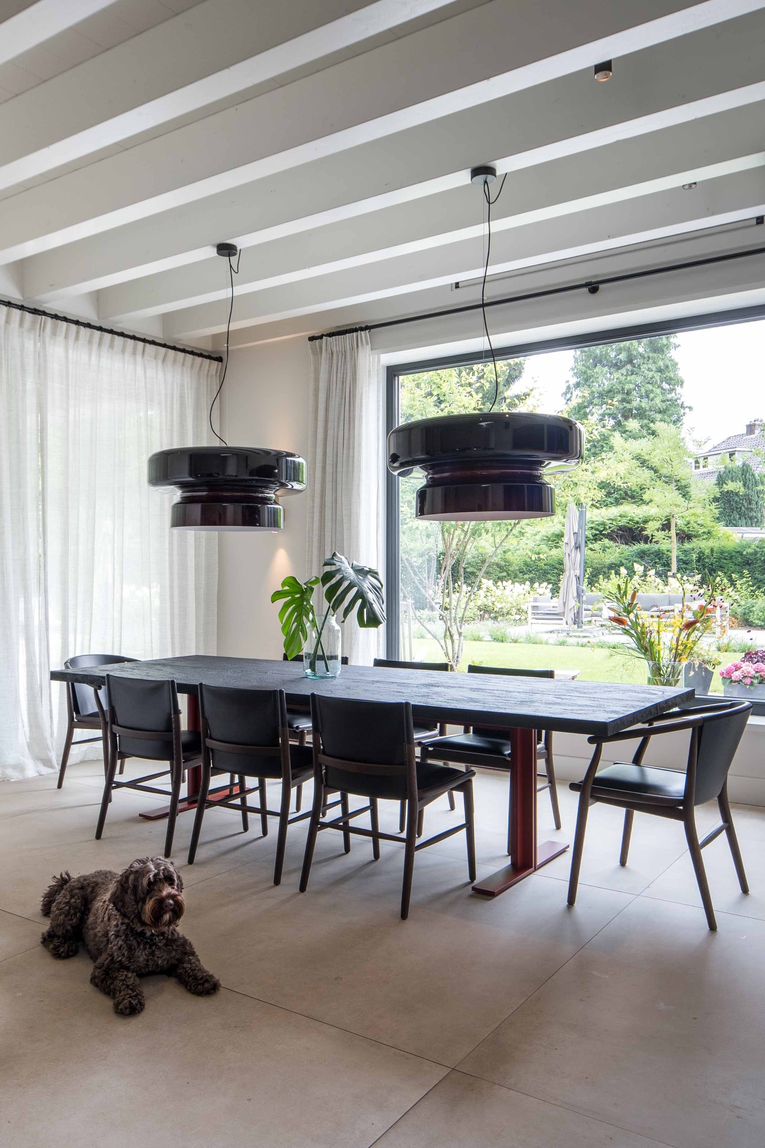 Eetkamer met donkere hangende lampen, zwarte stoelen en grote tuinramen
