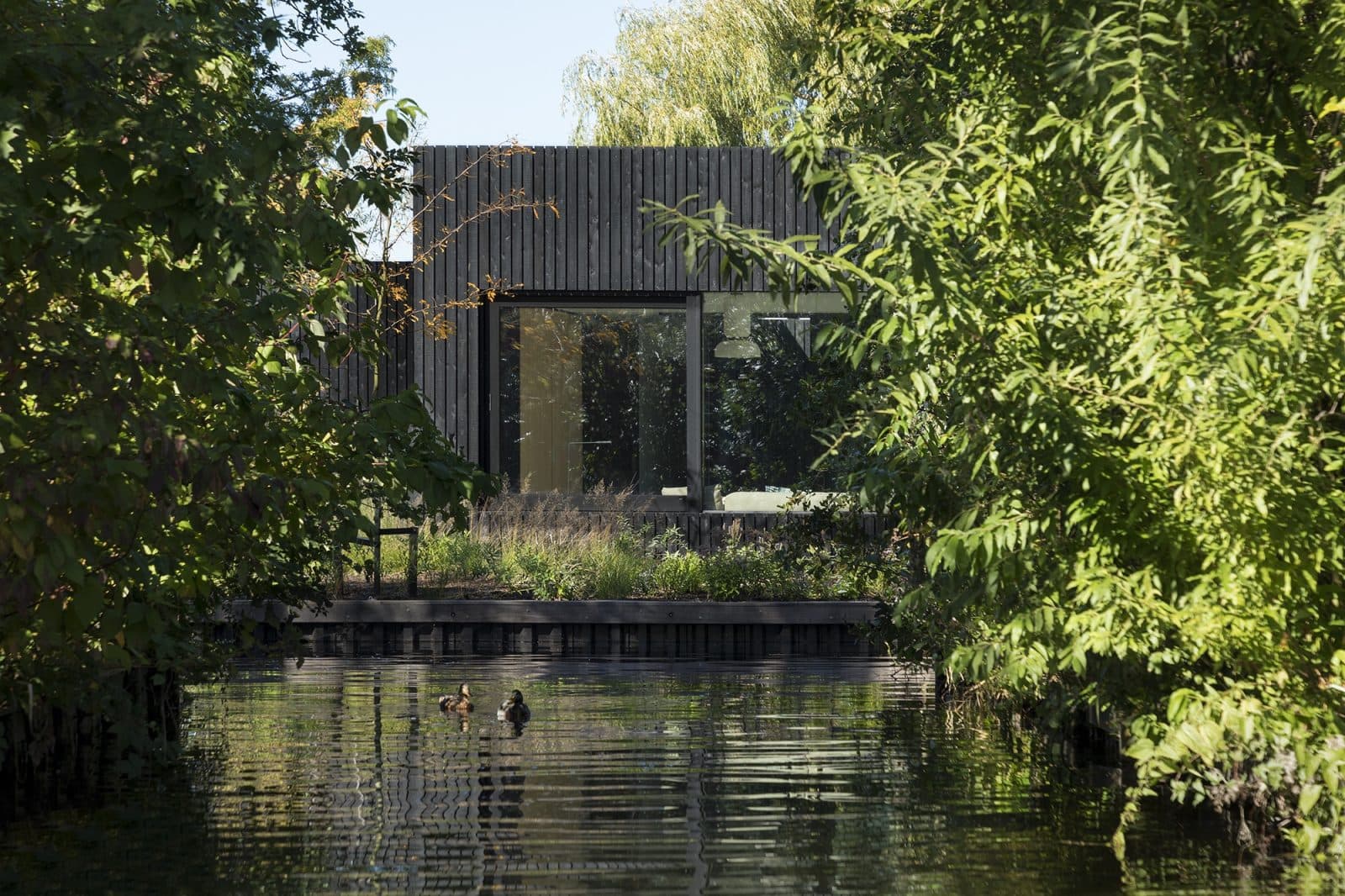 Zwart houten woonhuis aan water met dichte begroeiing aan oevers