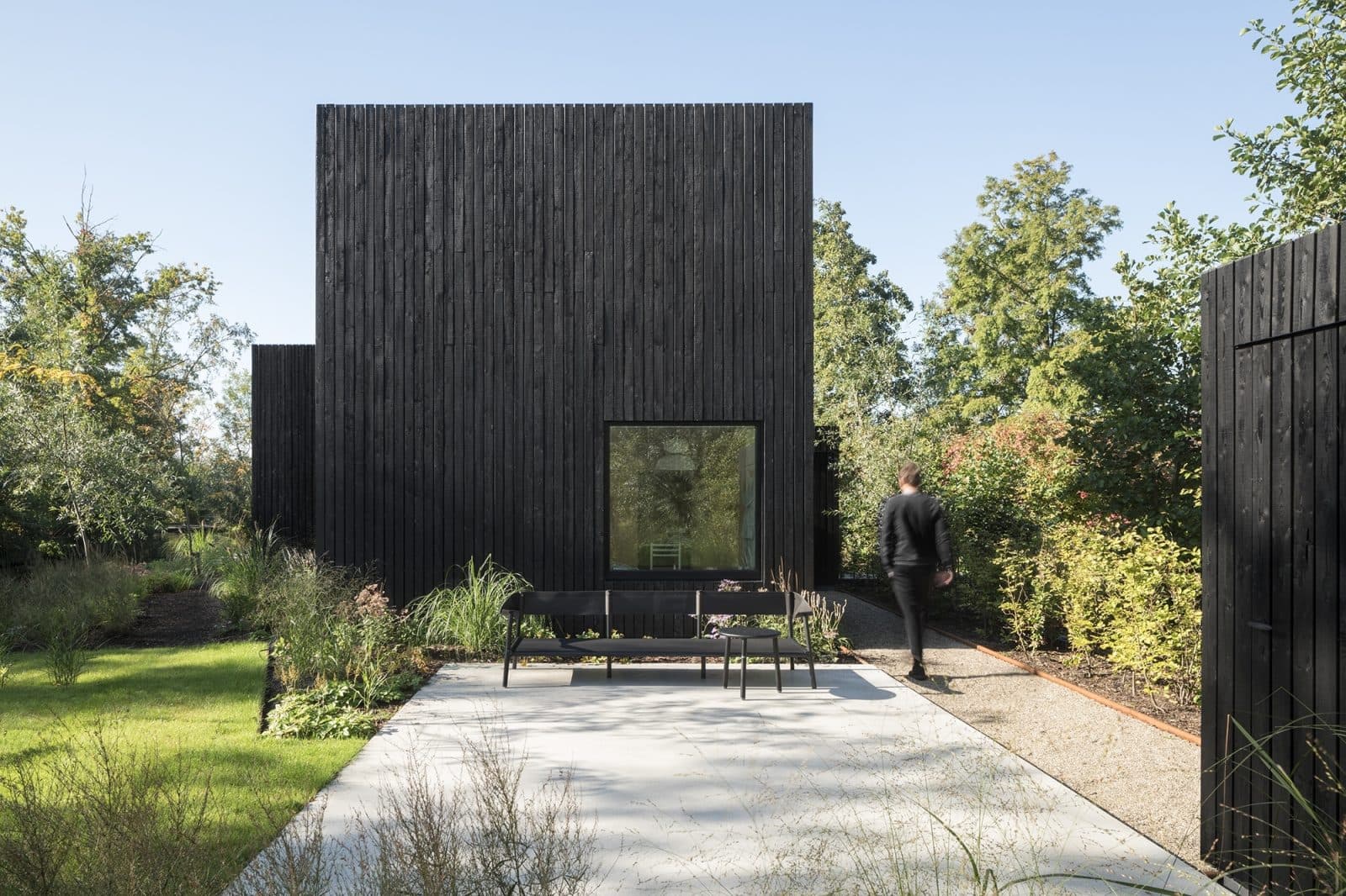 Zwart houten kubusvormig huis met brede beton inrit en natuurlijke plantenborder