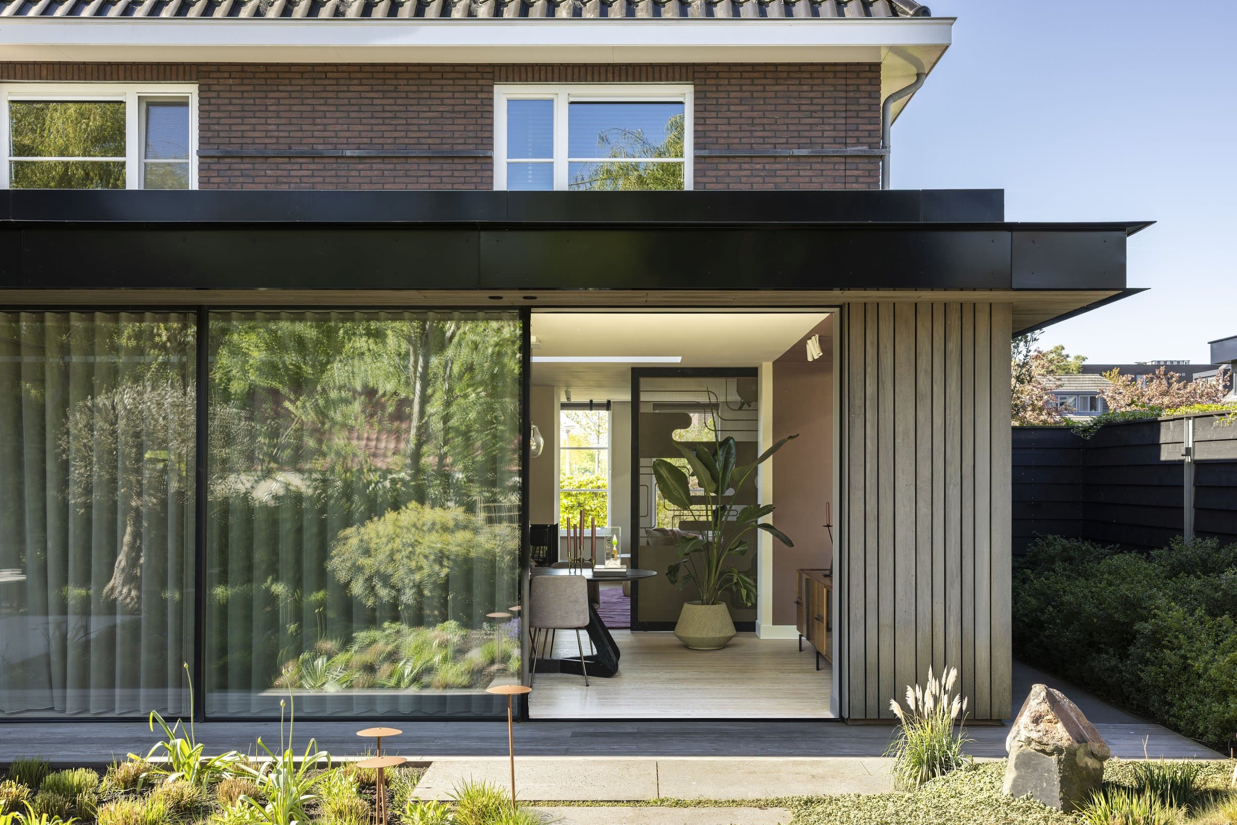 Bakstenen villa met witte schuifdeuren, zwarte zonwering en groene beplanting