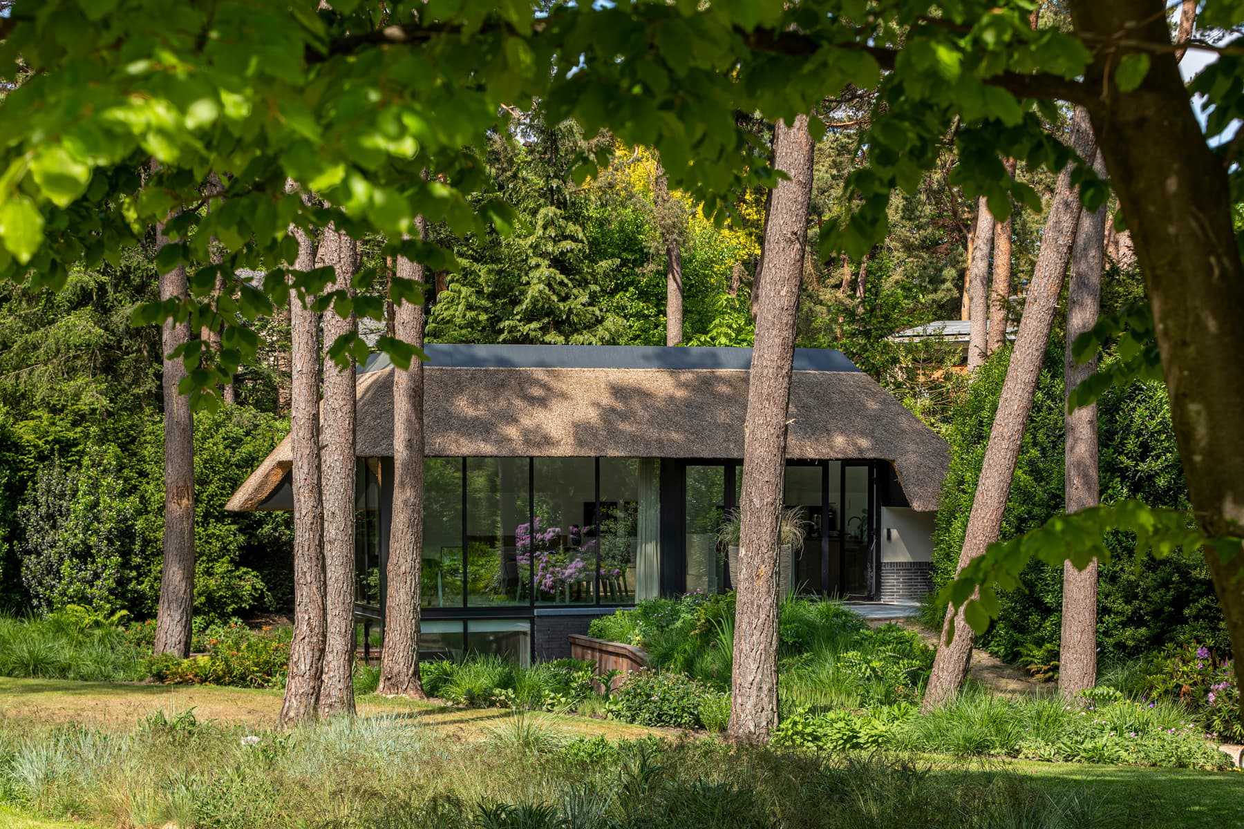 Tuinpaviljoen met rieten dak, grote raampartijen en veranda tussen hoge bomen