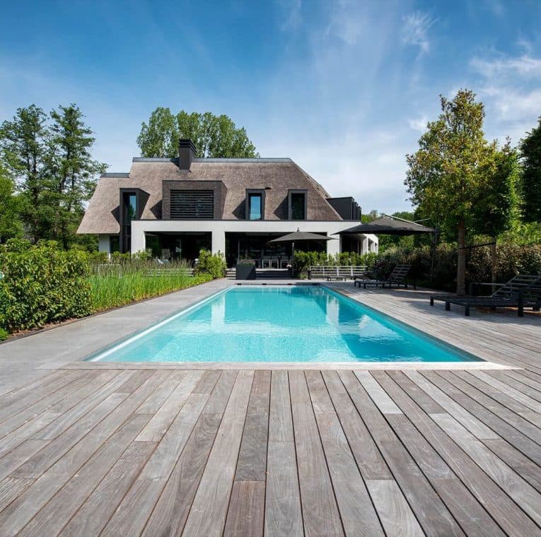 Zwembad met grijze houten terras, rietgedekte villa en tuin