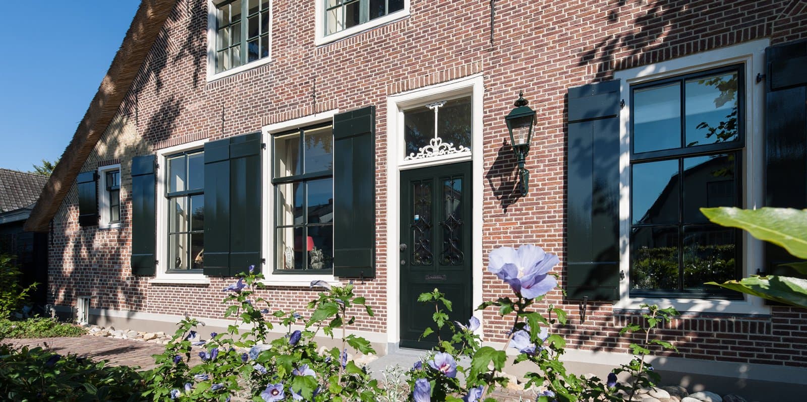 Voorgevel bakstenen huis met groene louvre-luiken en bloemen