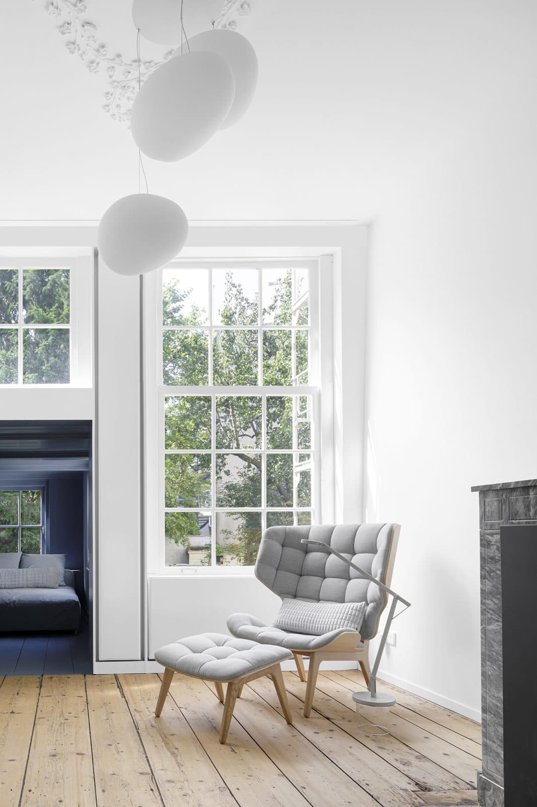 Woonkamer met grote ramen, witte hanglamp en grijze fauteuil met voetenbankje