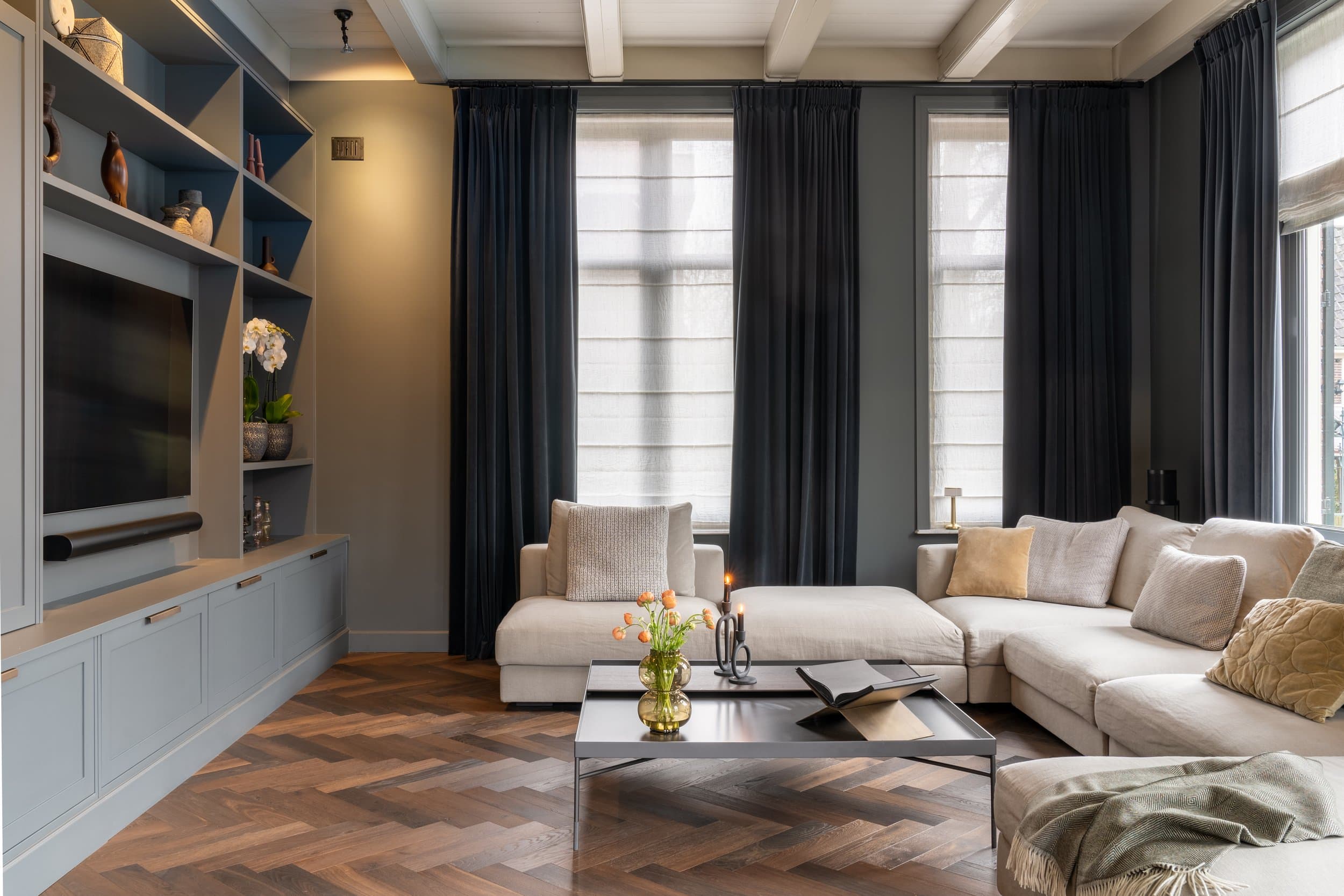 Woonkamer met grijze inbouwkasten, beige hoekbank en zwarte gordijnen